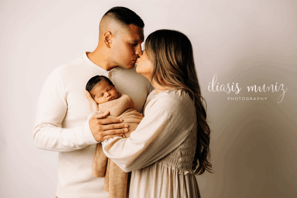 Newborn baby wrapped in a tan blanket being kissed by first-time parents during a professional studio photo session in Los Fresons, Texas.